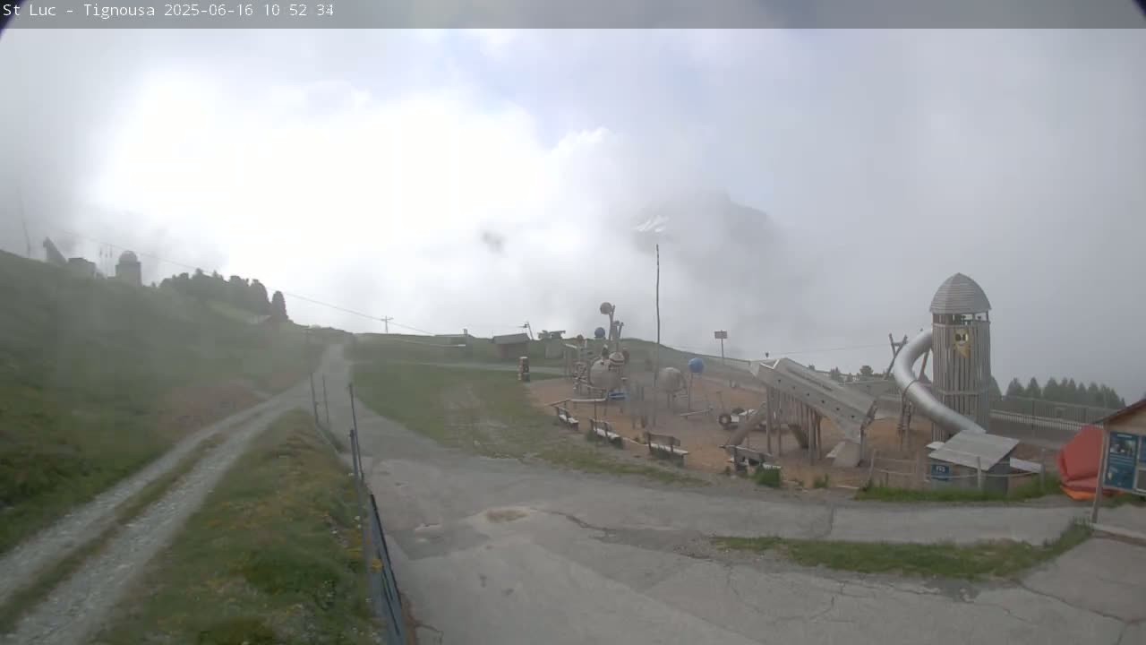 A playground with a large metal slide and wooden structures sits on a grassy hilltop shrouded in fog, with a gravel road leading down from it.