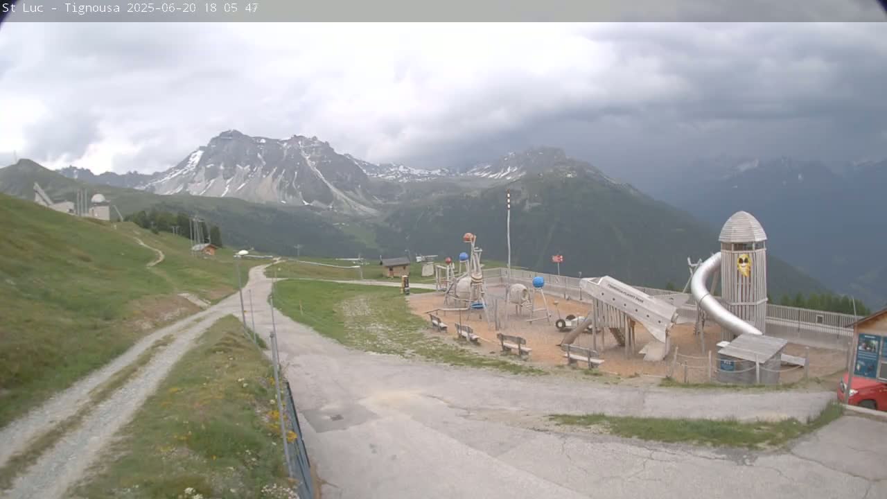 A playground sits on a grassy hill overlooking snow-capped mountains under a cloudy sky.