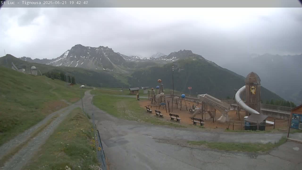 A playground sits in a mountainous area on an overcast day, with snow-capped peaks visible in the distance.
