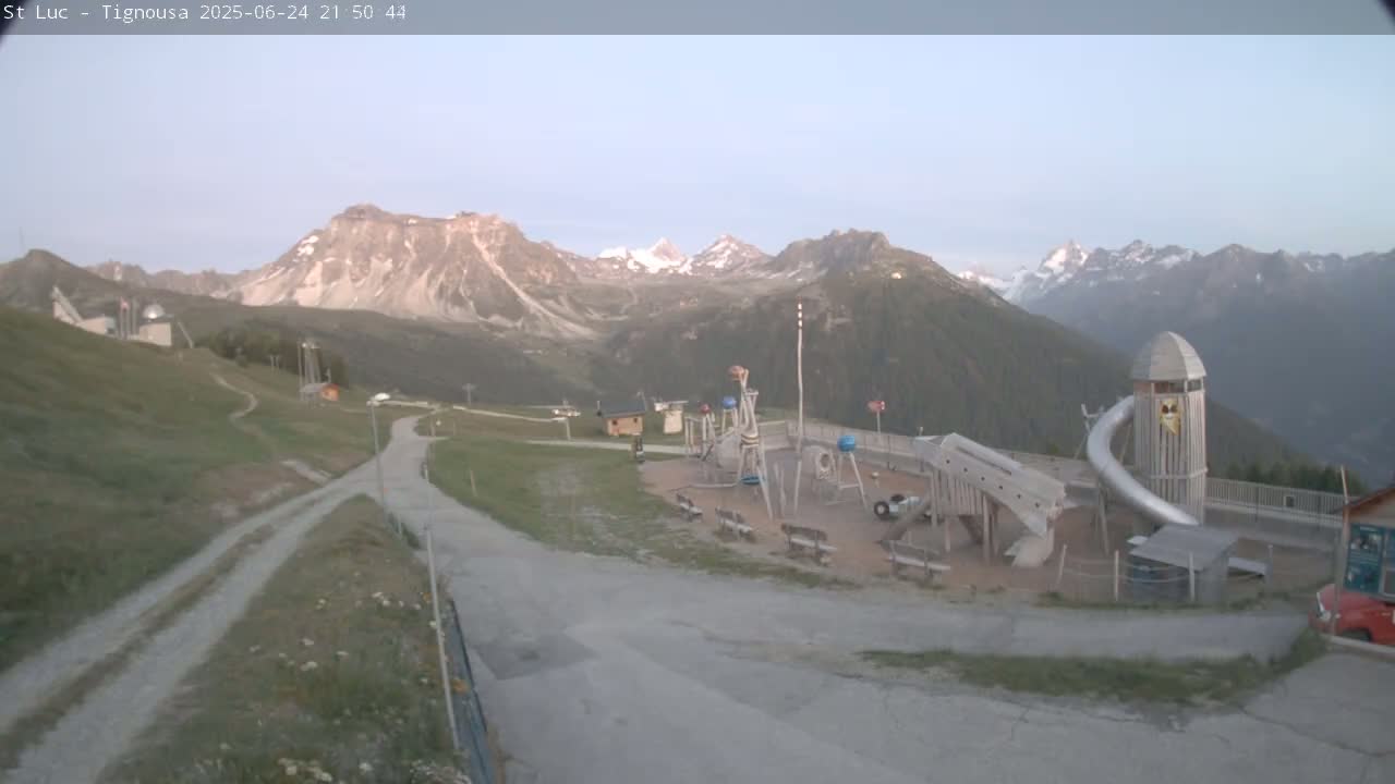 A playground with a long slide sits near a road winding through a grassy area, all set against a backdrop of mountains under a clear, twilight sky.