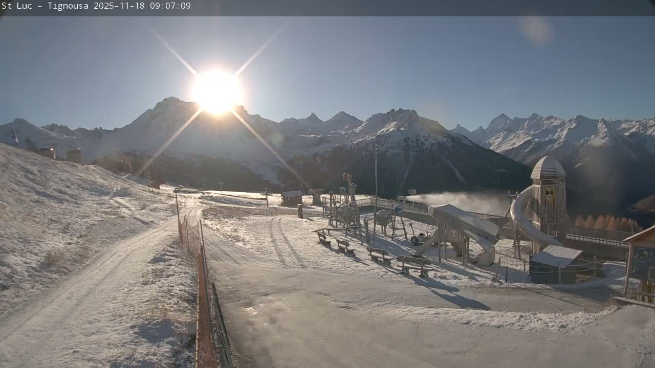 Under a clear blue sky on a bright winter morning, a snow-covered mountain scene features a playground and an active snow-making machine in the foreground, with the sun glinting over distant snow-capped peaks.