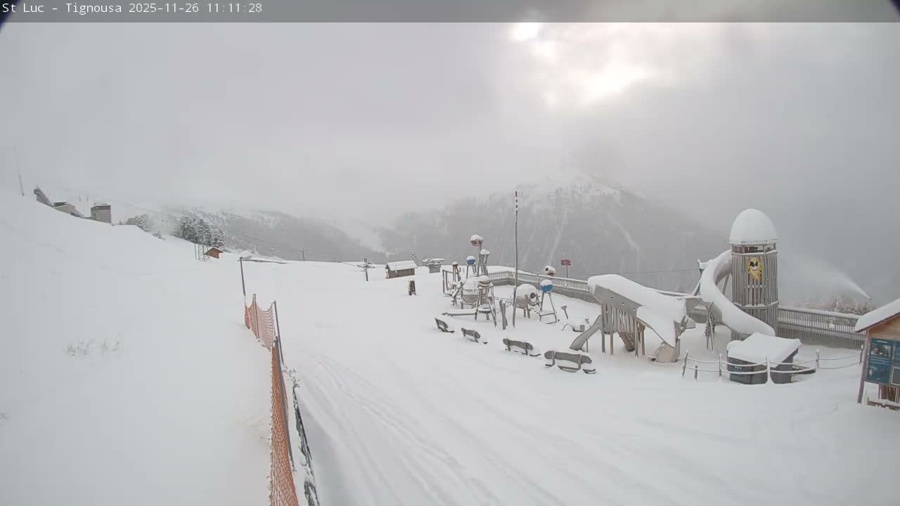 A snow-covered mountain resort features a playground, ski slope with orange netting, and an active snow cannon under an overcast and hazy winter sky.