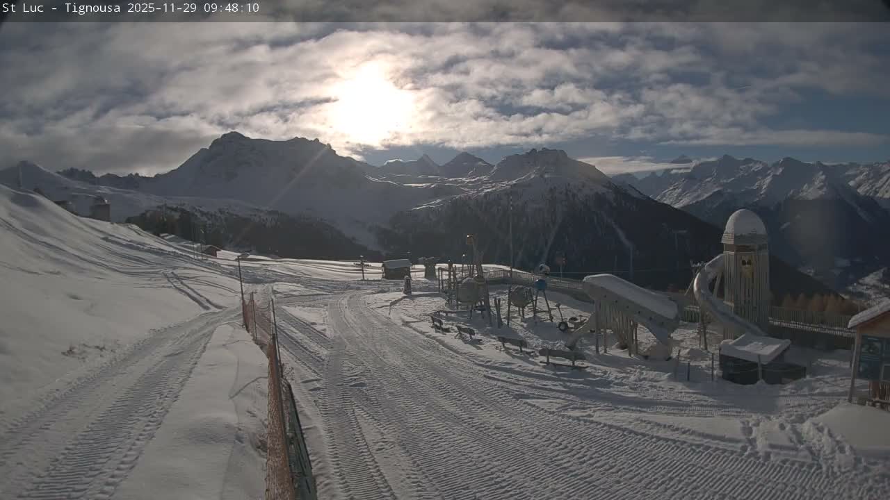 Snow-covered ski slopes and a playground are visible in a vast mountain landscape under a partly cloudy sky with a bright sun shining through.