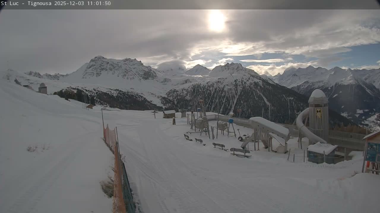 A snow-covered mountain ski area with a children's playground is depicted under a partly cloudy sky with sunlight breaking through.