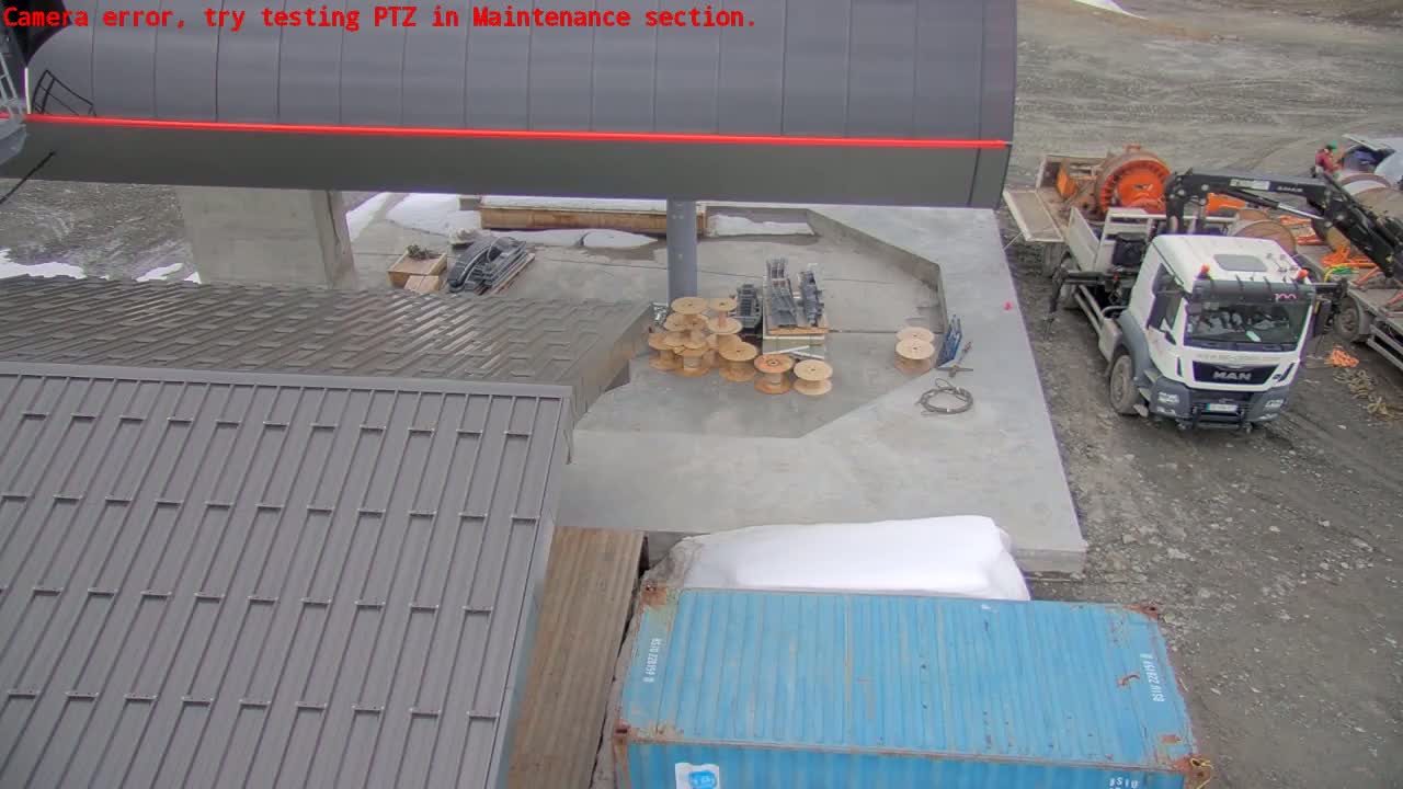 A high-angle view shows a construction site with a gray building, a blue shipping container, a truck, and several spools of cable on a concrete surface, partially covered in snow.