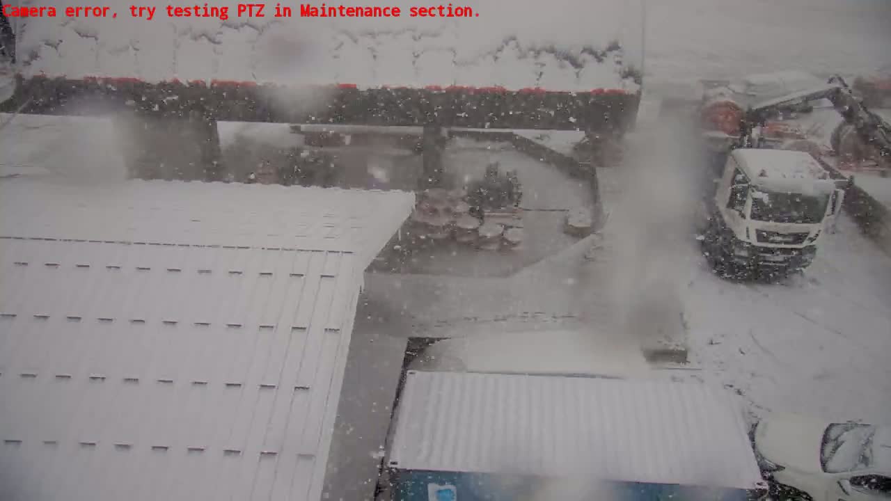 A snow-covered scene shows several vehicles, including a white truck and a car, in a heavy snowfall near a building with a snow-covered roof.