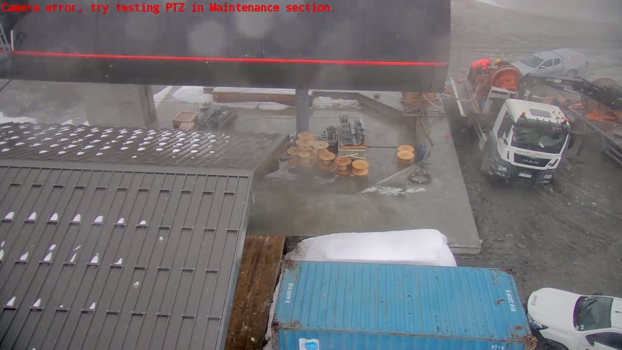 A snowy, overcast scene shows a construction area with various equipment, including a truck, a shipping container, and coils of cable, near a building with a gray roof.