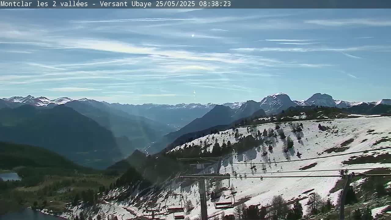 A mostly clear, sunny day reveals a snow-dusted mountainside with a chairlift, overlooking a vast valley and distant, snow-capped mountain range.