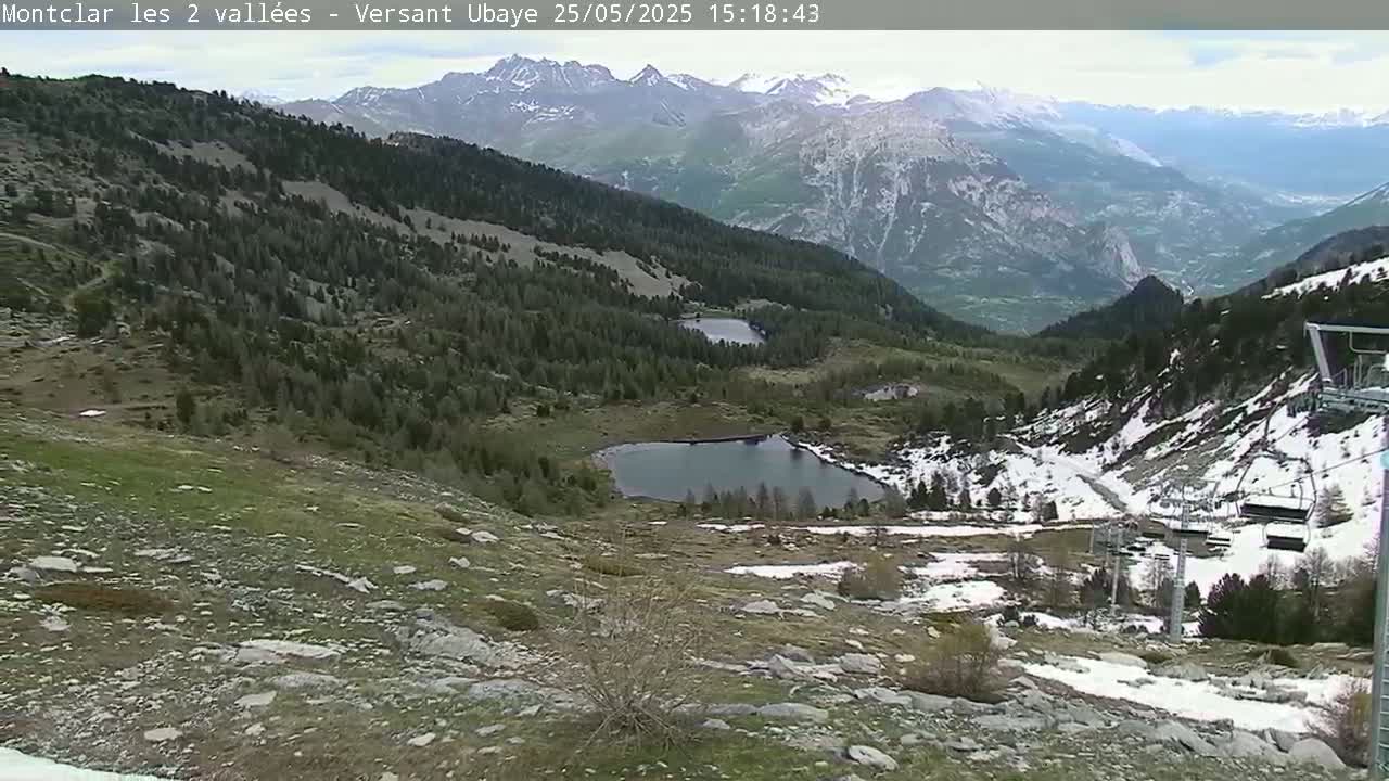 A mountainside view shows two small alpine lakes nestled among green and brown hills, with patches of snow remaining on the ground and distant mountains under a partly cloudy sky.