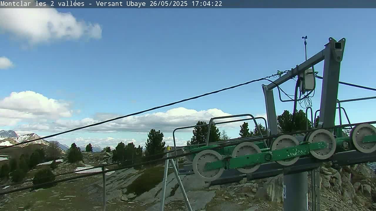 A mostly clear blue sky with some fluffy white clouds overlooks a mountaintop ski lift with several large pulleys and a rocky landscape.