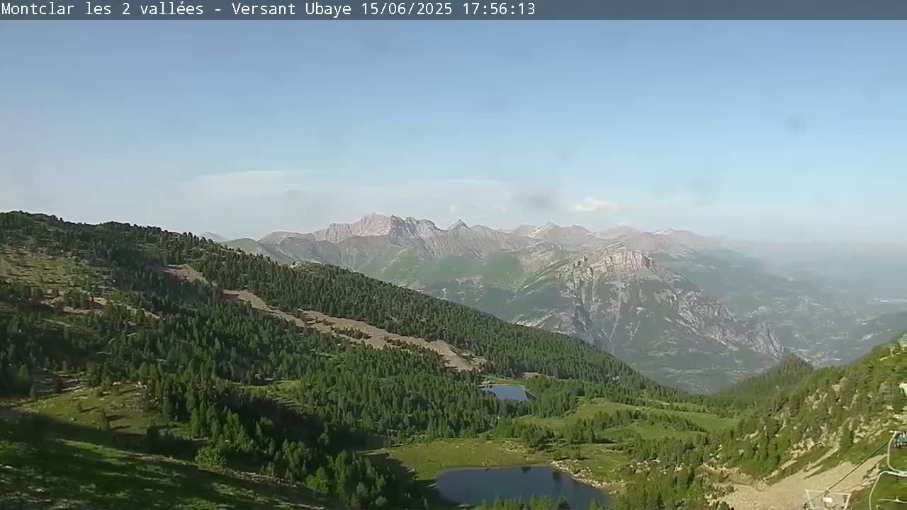 A panoramic view of a mountainous region shows a lush green landscape with two small lakes, under a clear, sunny sky.