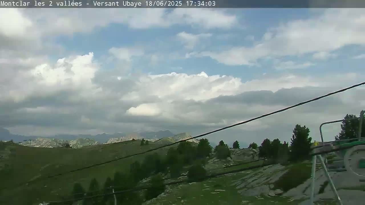 A partly cloudy sky overlooks a mountainous landscape with green hills, sparse trees, and rocky areas, partially obscured by chairlift cables.