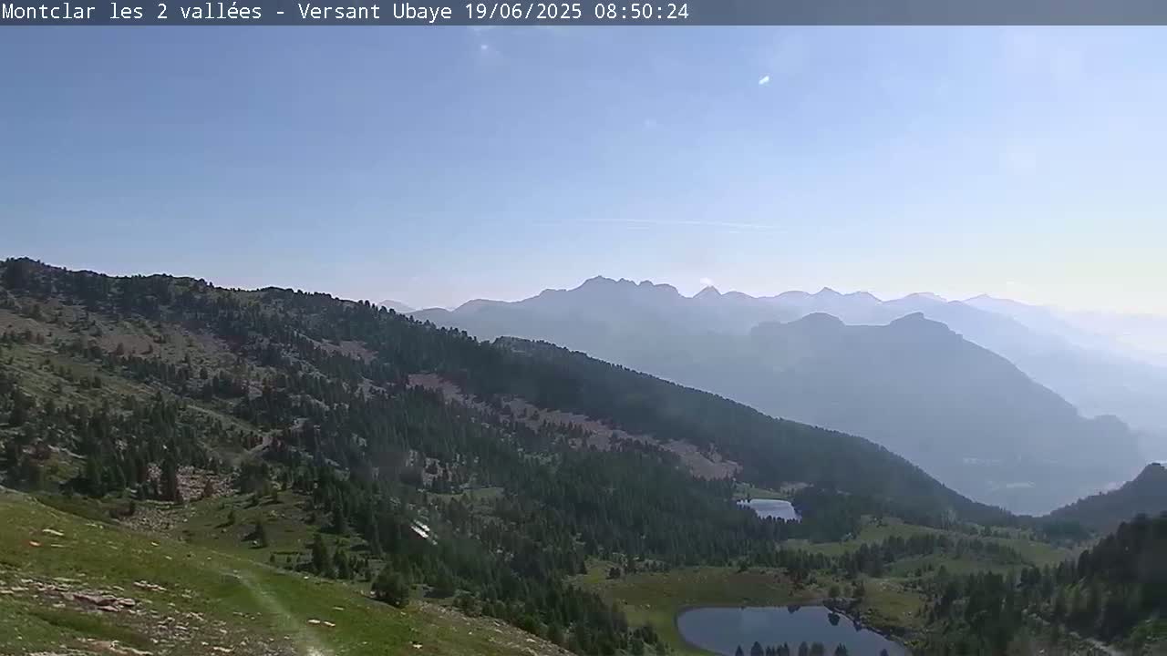 A hazy, sunny day reveals a mountainous landscape with coniferous forests, grassy meadows, and several small lakes nestled in the valleys.