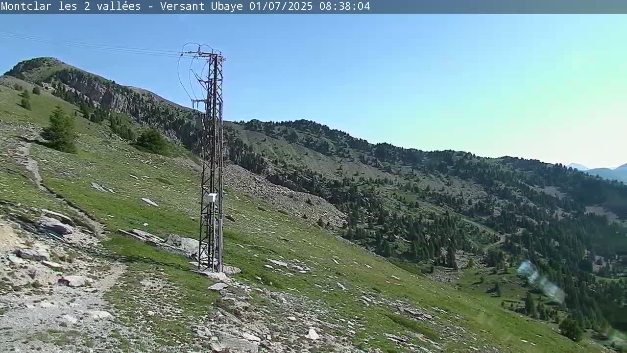 A rocky, grassy mountain slope with scattered trees under a clear blue sky is shown, and a tall metal utility pole stands near the foreground.