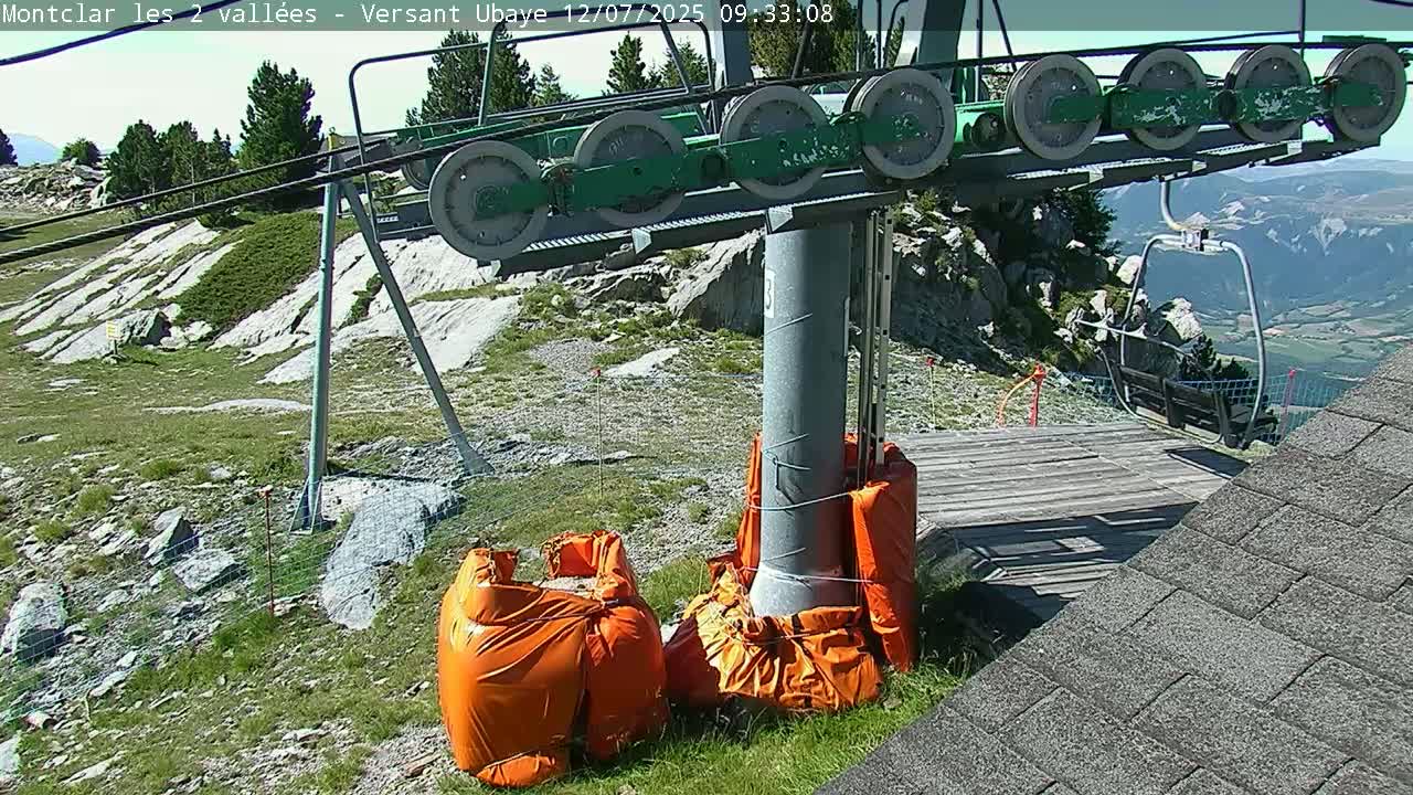 A chairlift terminal on a sunny day, with orange protective coverings around its base, sits on a rocky hillside overlooking a valley.