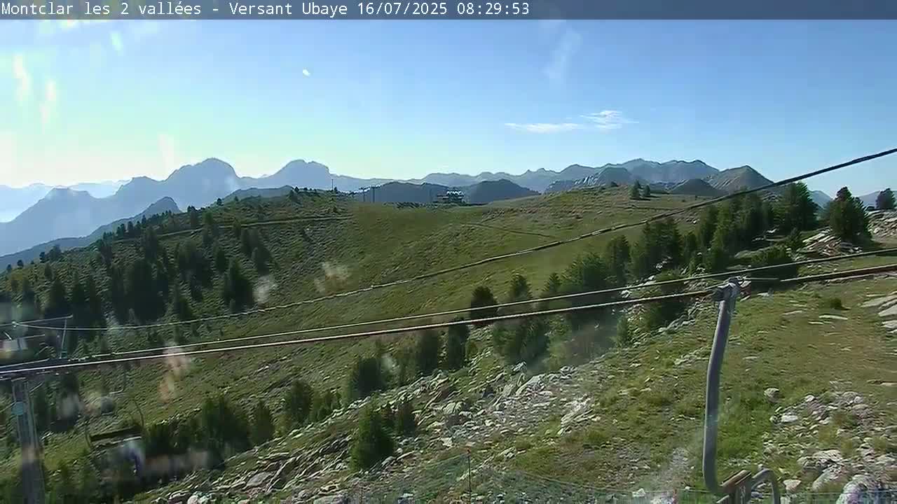 A mostly sunny view of a mountainous landscape with green hills and sparse trees, seen from a chairlift.