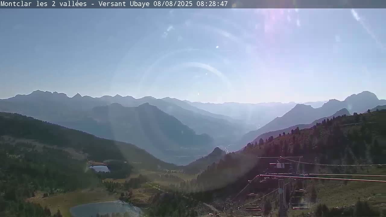A sunny, hazy day reveals a mountainous landscape with a valley below, featuring a small lake and a chairlift in the foreground.