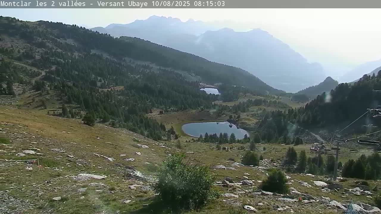 A hazy mountain valley scene features two small lakes nestled among evergreens, with rocky, grassy slopes leading down to a chairlift in the foreground.