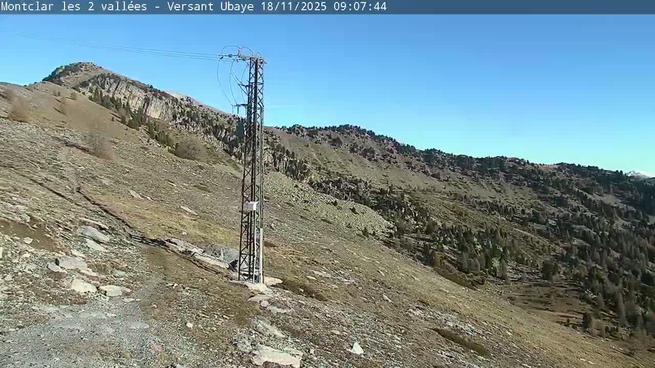 A clear, sunny day reveals a rugged mountain landscape with a prominent power line pylon standing on a rocky, sparsely vegetated slope under a bright blue sky.
