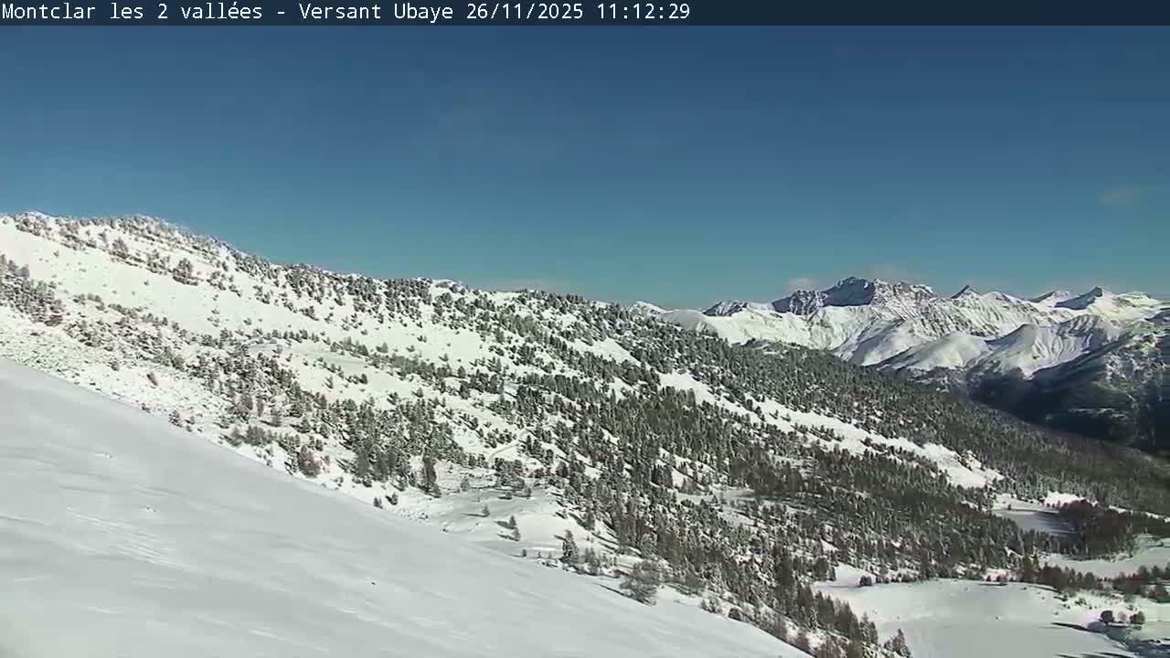A vast, snow-covered mountain landscape unfolds under a clear blue sky, with a smooth snowy slope in the foreground leading up to forested hills and jagged peaks in the distance.
