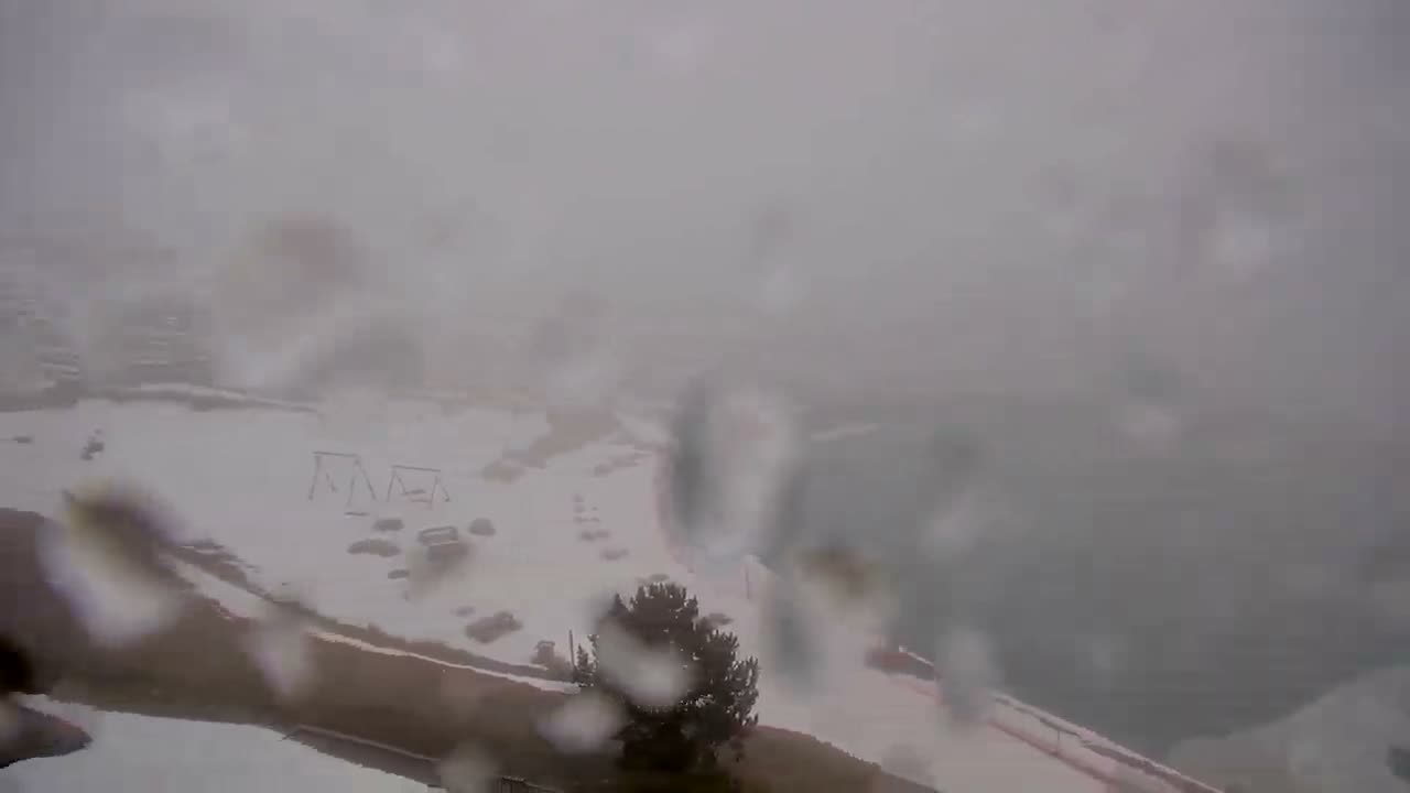 A very blurry, snow-covered outdoor landscape is seen through a precipitation-covered camera lens, indicating heavy snowfall or dense fog with extremely low visibility.