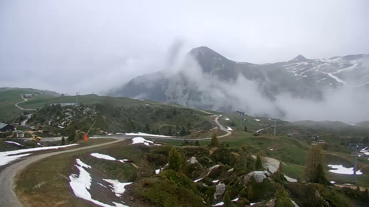 A foggy mountain landscape shows a winding road curving through green hills speckled with patches of snow, with ski lifts and buildings visible in the distance.