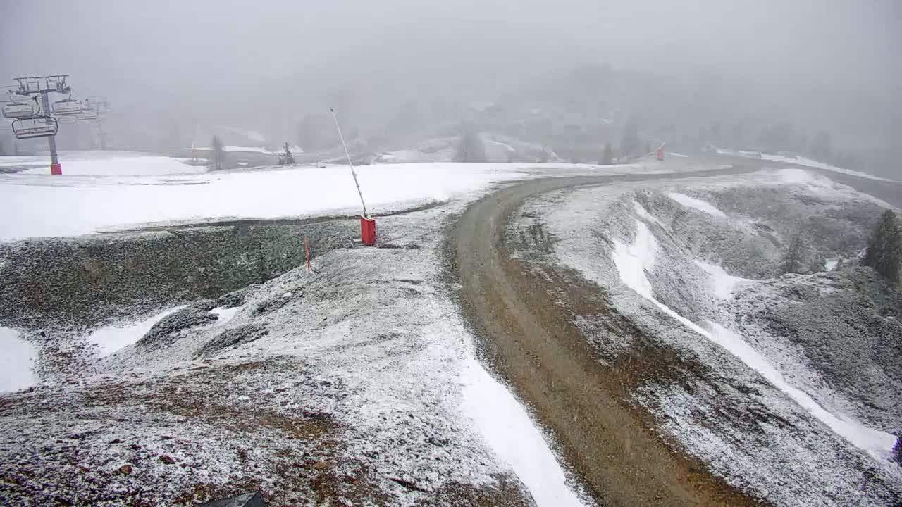 A snow-covered, hilly landscape with a dirt road winding through it, featuring a ski lift in the background under foggy conditions.
