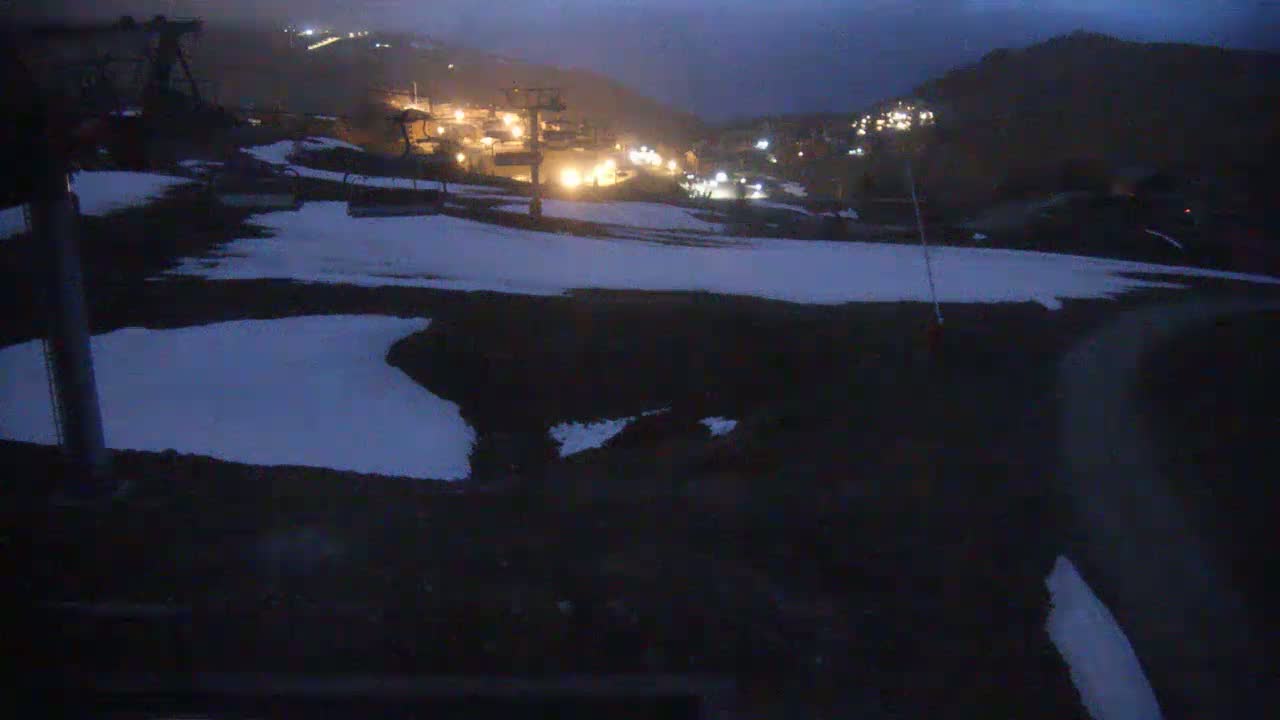 A nighttime view of a snow-dusted ski resort with sparsely lit buildings and ski lifts on a dark, cloudy night.