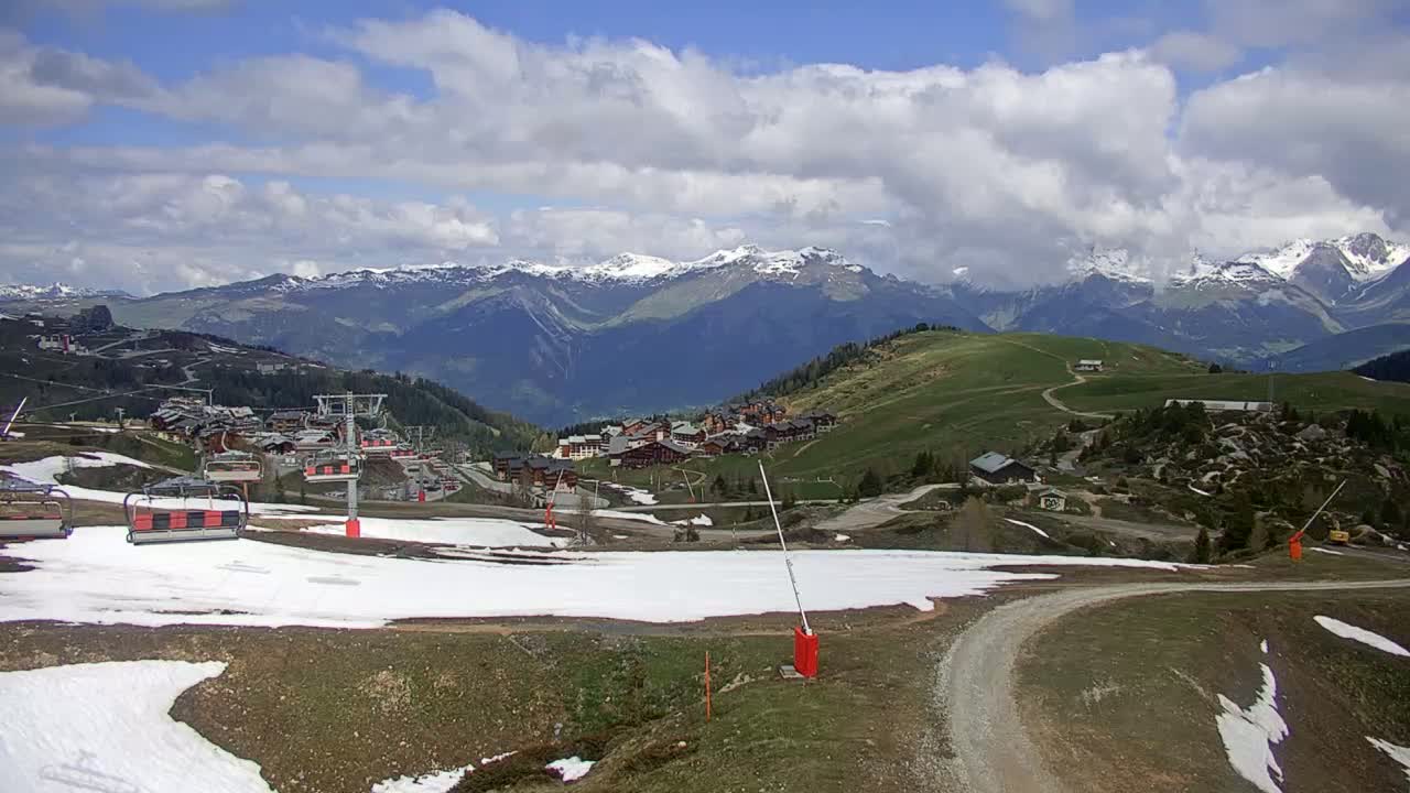 A partly cloudy day reveals a mountain village nestled in a valley, with patches of snow remaining on the ground near a chairlift and winding dirt road.