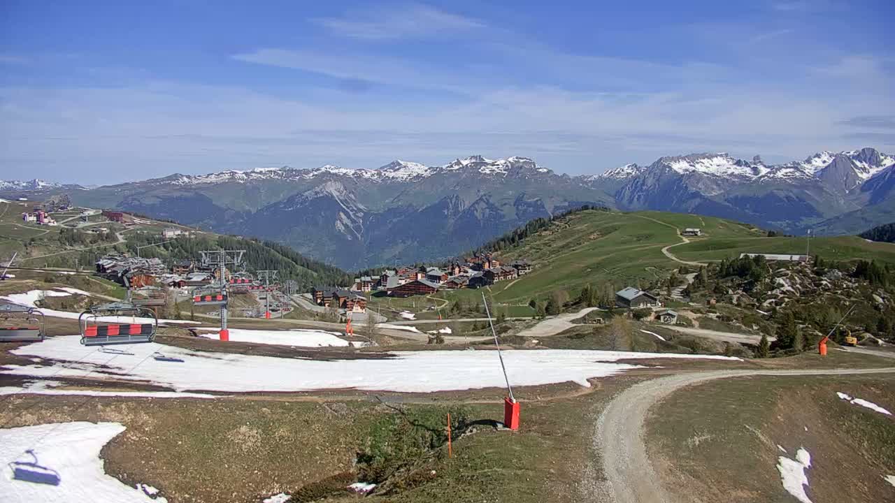 A sunny mountain landscape shows a ski resort with chairlifts, patchy snow, and a dirt road winding through green hills toward snow-capped mountains in the distance.