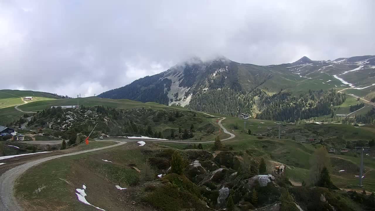 A winding road leads through a grassy mountain landscape with patches of snow under a cloudy sky, towards a peak shrouded in mist.