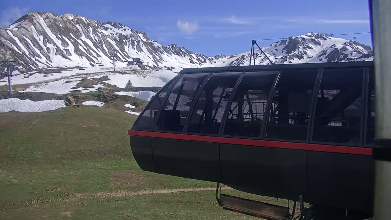 A gondola car with a red stripe hangs suspended above a grassy hillside dotted with patches of snow, with snow-capped mountains in the background under a mostly sunny sky.