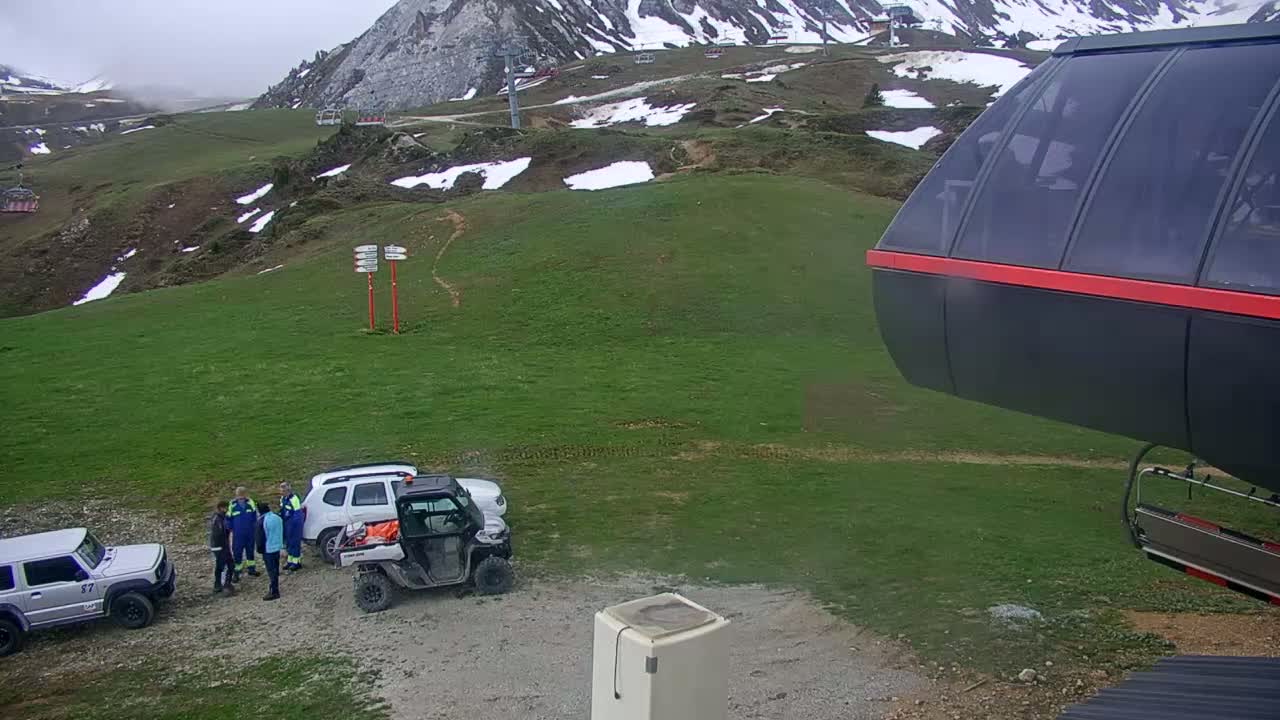 A grassy mountainside with patches of snow, several vehicles, a group of people, and a portion of a chairlift car are visible under overcast skies.
