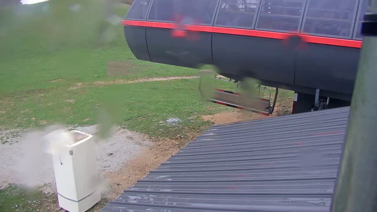 A rain-streaked view shows a chairlift gondola and a section of corrugated metal, with a grassy area and a white appliance in the background.