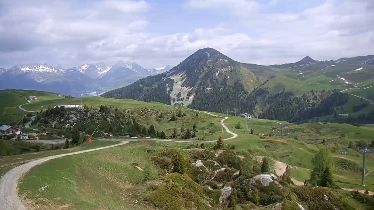 A winding dirt road snakes through rolling green hills and past rocky outcrops, with snow-capped mountains in the background under a partly cloudy sky.