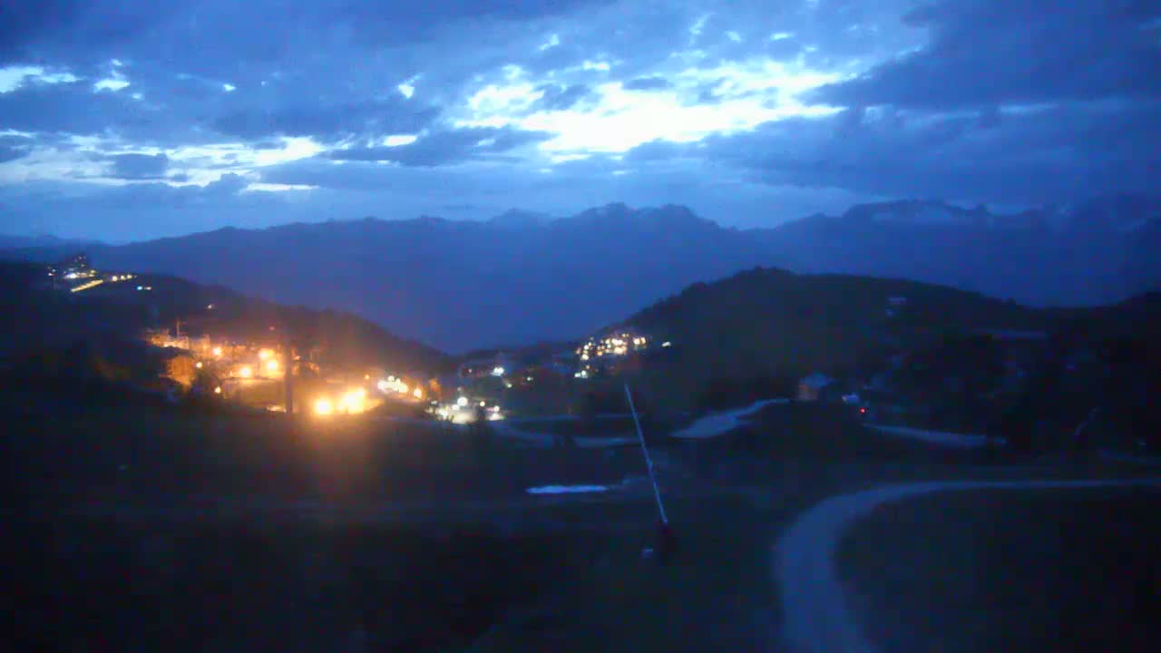 A mountain village at night is illuminated by scattered lights against a backdrop of dark, cloudy mountains under a twilight sky.