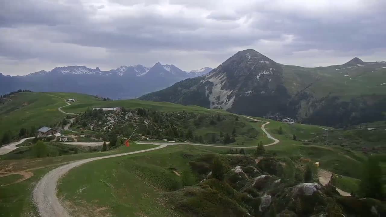 A winding dirt road meanders through a mountainous landscape under a cloudy sky, passing small buildings and rocky outcrops.