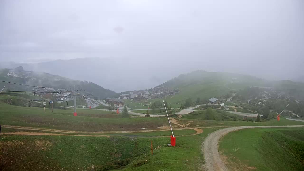 A foggy mountain village is viewed from a hillside, with a chairlift and ski resort infrastructure visible in the mid-ground.
