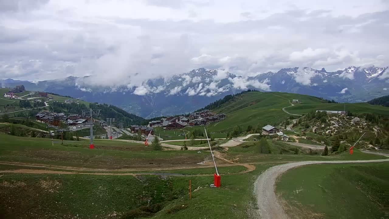 A mountain village nestled in a green valley, under a cloudy sky with snow-capped mountains in the background.