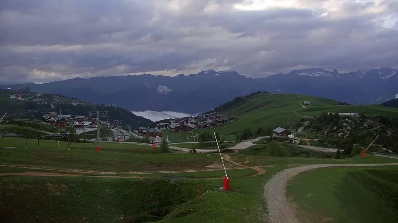 A mountain village nestled in a valley, under a cloudy sky, is surrounded by green hills and distant, snow-capped mountains.