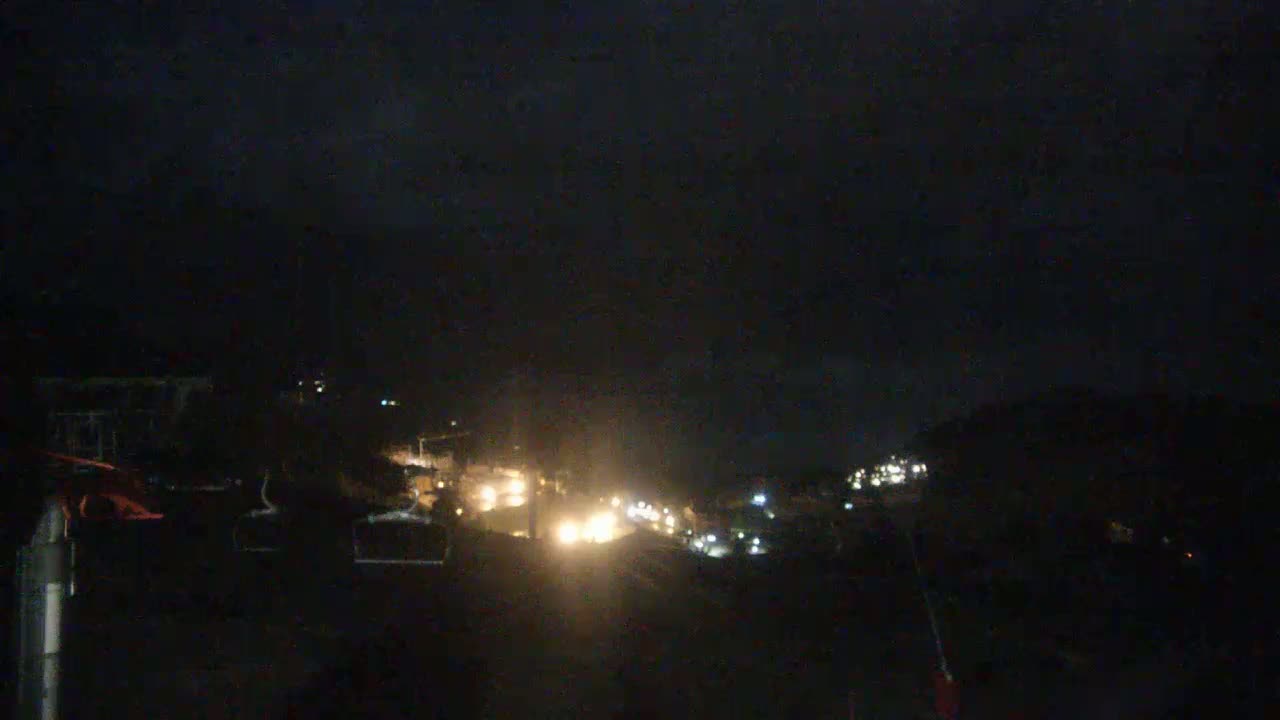 A nighttime long shot of a dimly lit road winding through a dark landscape, with a few lights visible along the roadside and in the distance.