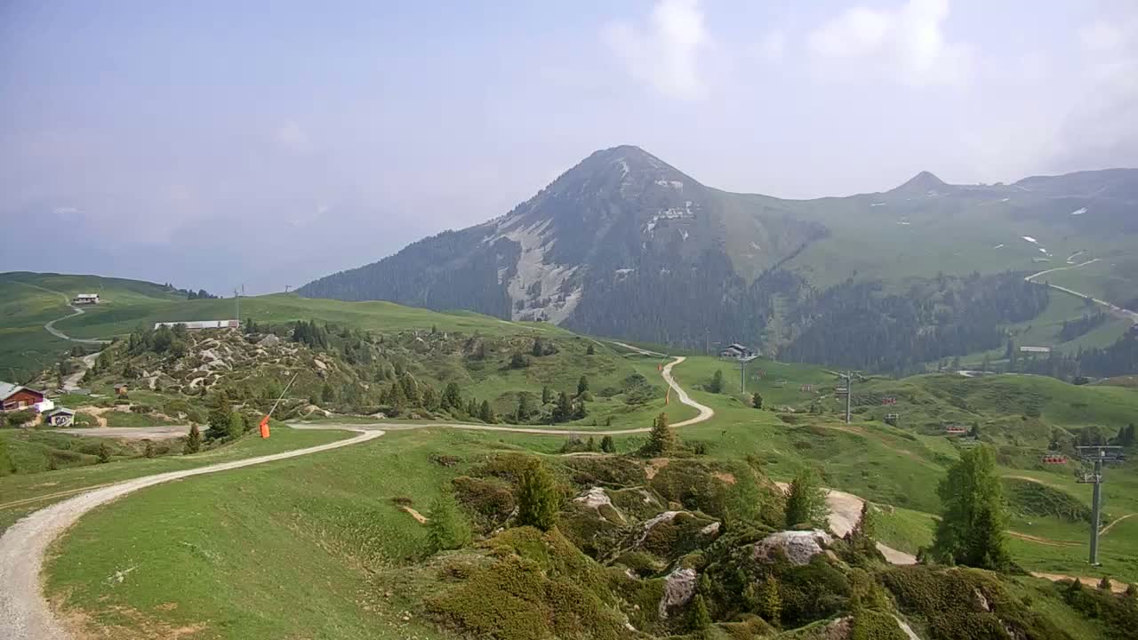 A winding dirt road traverses rolling green hills dotted with rocks and sparse trees, leading toward a prominent mountain under a mostly clear, hazy sky.