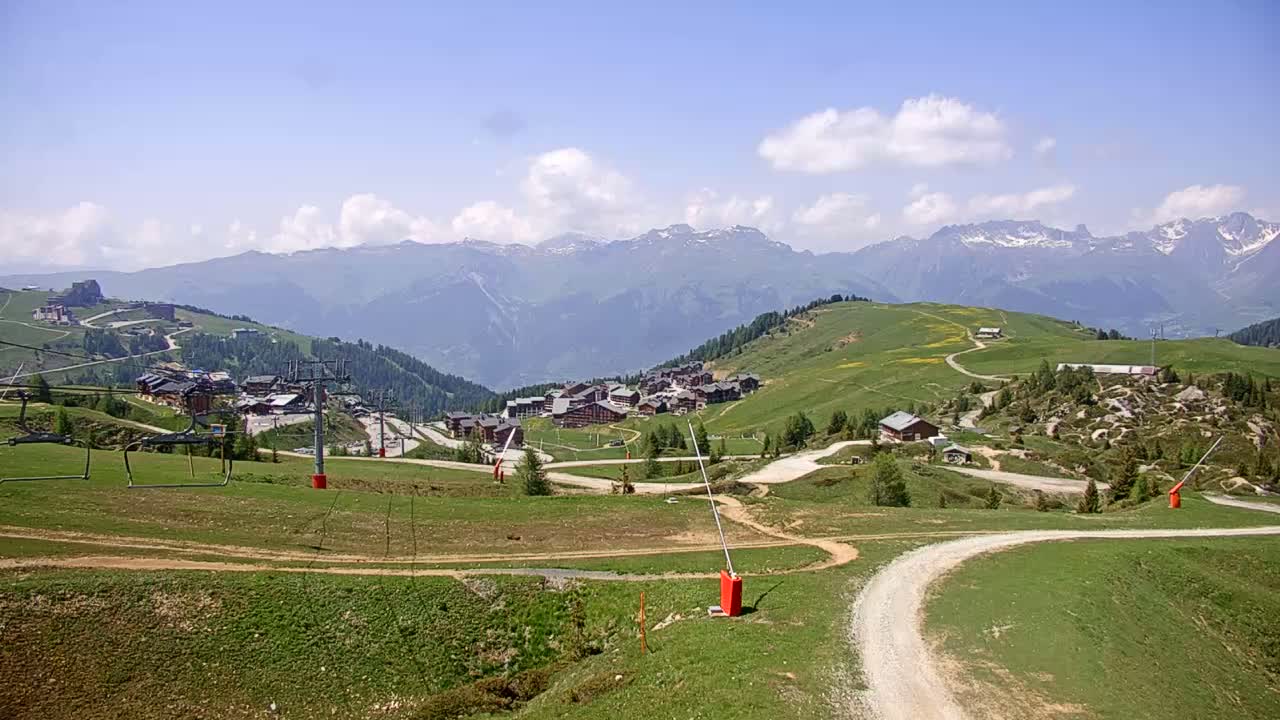 A mountain village nestled in a green valley under a partly cloudy blue sky, with ski lifts and winding roads visible.