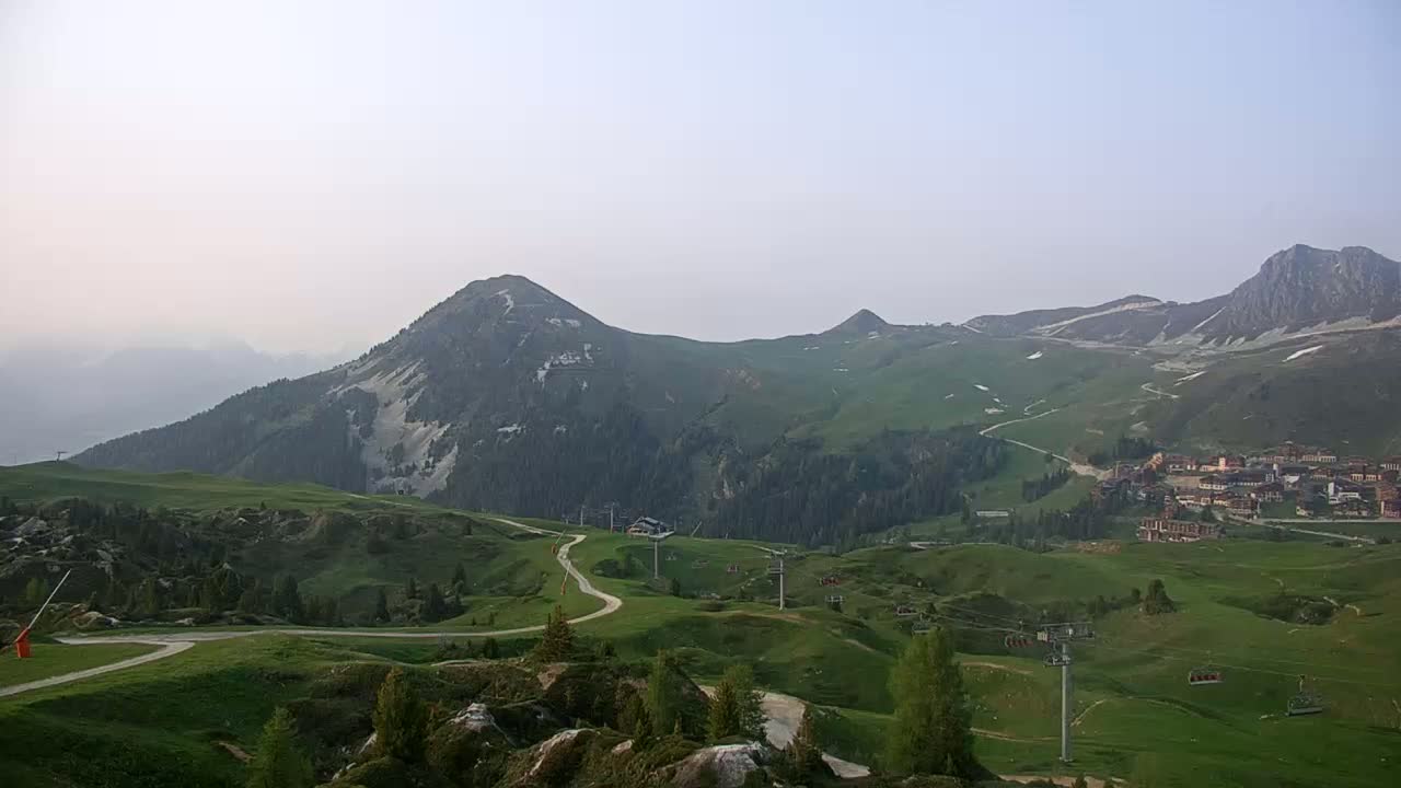 Rolling green hills and mountains under a hazy sky, with ski lifts and a small village visible in the distance.