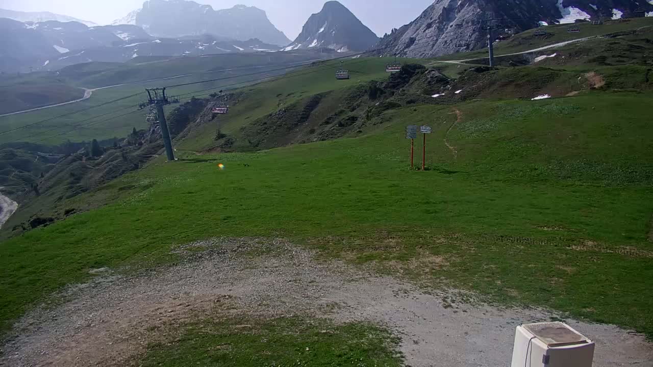 A grassy mountaintop area with a chairlift, some small buildings, and distant snow-capped mountains under a clear, sunny sky.