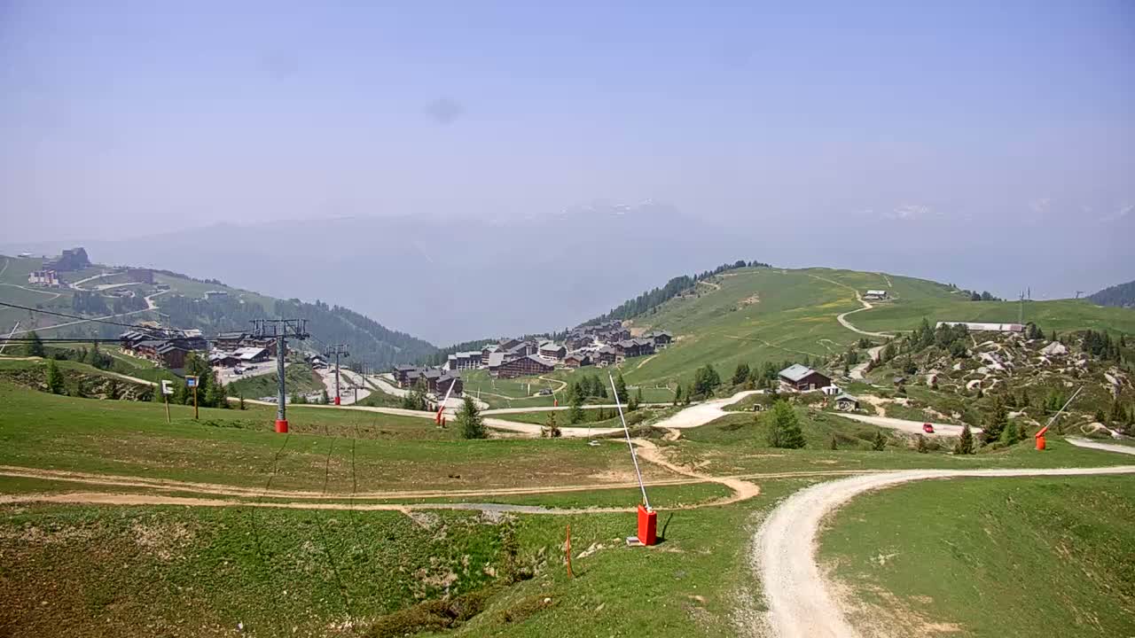 A hazy mountain village with ski lift infrastructure is visible on a grassy hillside under a clear sky.