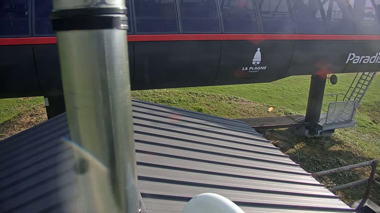 A metal pole is in the foreground, partially obscuring a view of a dark-colored gondola lift against a sunny, grassy background.