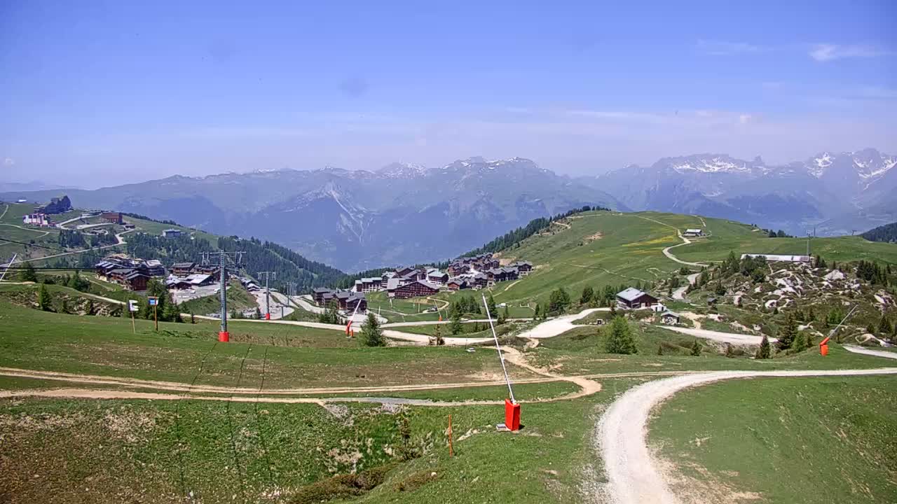 A mountain village nestled in a valley, under a clear blue sky, is surrounded by green hills and distant snow-capped mountains.