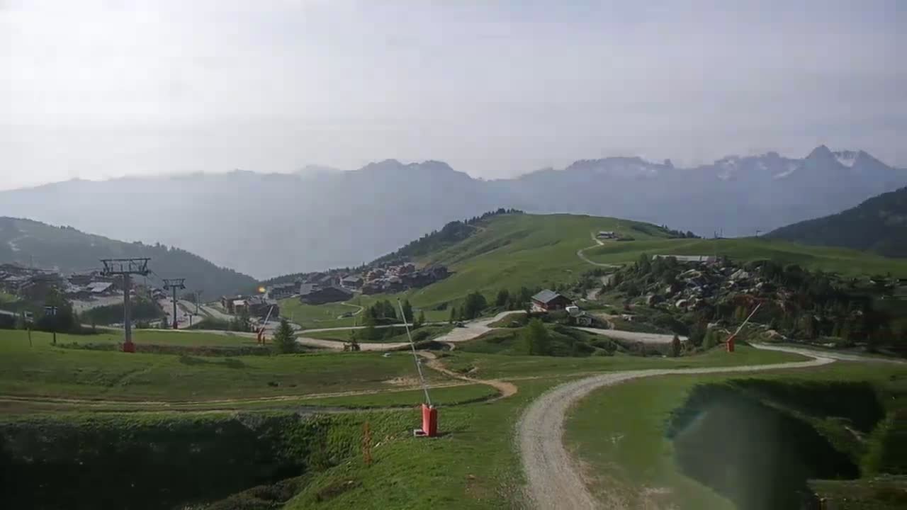 A hazy mountain landscape shows a grassy hillside with a winding dirt road, ski lifts, and small buildings nestled among the green slopes, mountains in the background.