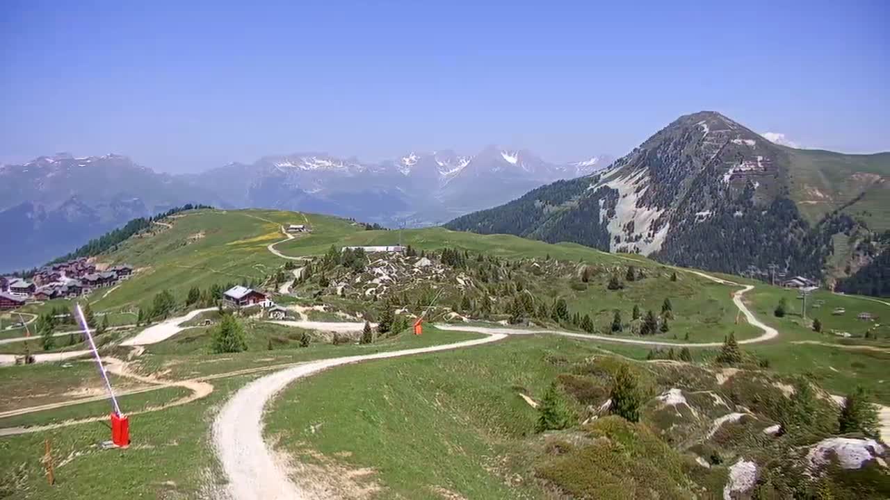 A winding dirt road meanders through a green mountain landscape under a clear blue sky, with snow-capped mountains in the distance.
